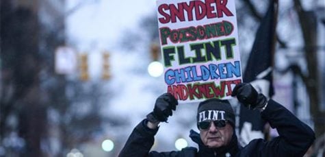 Mike Ahrens of Muskegon, Mich., poses for photo with his sign about Flint's water crisis Monday, Jan. 18, 2016, in Ann Arbor, Mich. Michigan Gov. Rick Snyder responded Monday to criticism from presidential candidate Hillary Clinton during the Democratic debates for his handling of Flint's water emergency, saying Clinton is making it a political issue. (Junfu Han/The Ann Arbor News via AP) LOCAL TELEVISION OUT; LOCAL INTERNET OUT; MANDATORY CREDIT