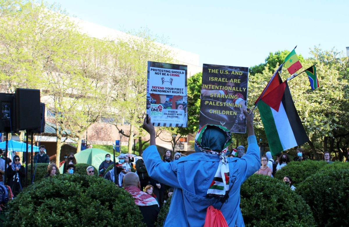 Un manifestante sostiene dos carteles pro-palestinos, mientras los compañeros manifestantes cantan en solidaridad con Gaza el martes 30 de abril de 2024. El Taller de Desinversión del campamento se llevó a cabo entre las 4:30 y las 6:00 p.m. del martes.